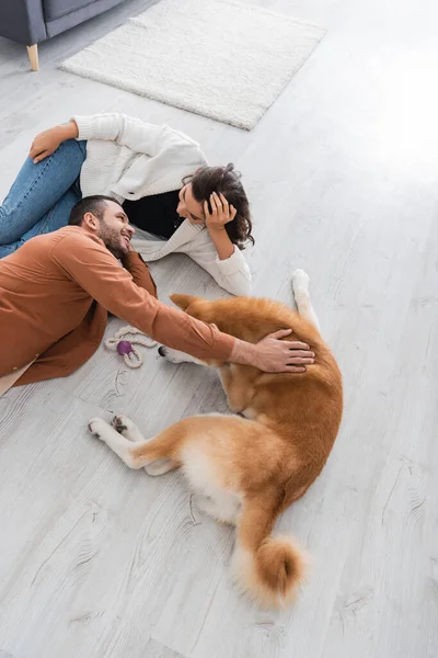 Top view of joyful young couple looking at each other while lying of floor with akita inu dog — Stock Photo