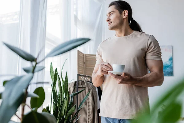 Man with long hair holding cup of coffee and saucer near green plants — Stock Photo