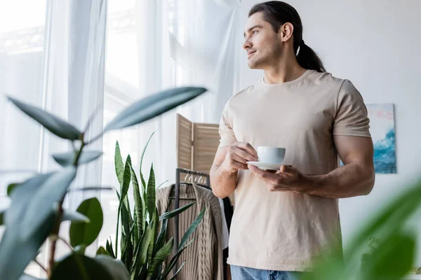 Homme aux cheveux longs tenant tasse de café et soucoupe près de plantes vertes — Photo de stock