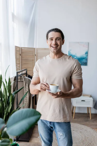 Homme gai tenant tasse de café et soucoupe près de plantes vertes — Photo de stock