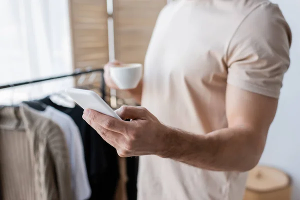 Vista recortada del hombre sosteniendo la taza de café y teléfono inteligente - foto de stock