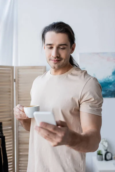 Hombre en camiseta sosteniendo la taza de café y el uso de teléfono inteligente - foto de stock