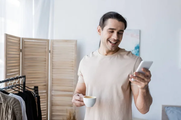 Homme heureux en t-shirt tenant tasse de café et en utilisant smartphone — Photo de stock