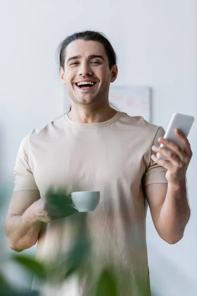 Homem feliz em t-shirt segurando xícara de café e smartphone — Fotografia de Stock