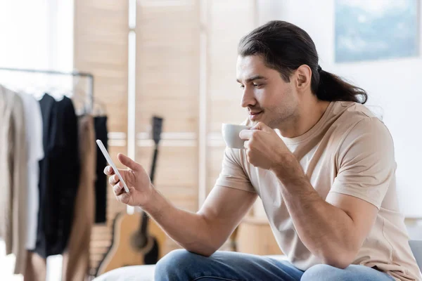 Homme aux cheveux longs tenant une tasse de café et utilisant un téléphone mobile — Photo de stock