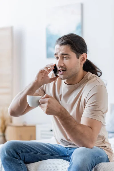 Мужчина с длинными волосами держит чашку кофе и разговаривает по мобильному телефону — стоковое фото