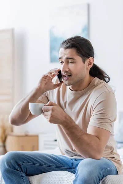 Man with long hair holding cup of coffee and talking on mobile phone — Stock Photo