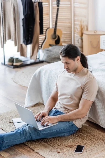 Чоловік з довгим волоссям, використовуючи ноутбук, сидячи на підлозі в спальні — стокове фото