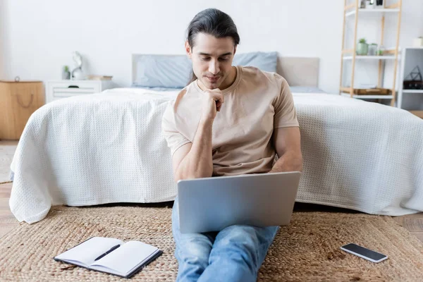 Freelancer using laptop near smartphone with blank screen and notebook in bedroom — Stock Photo