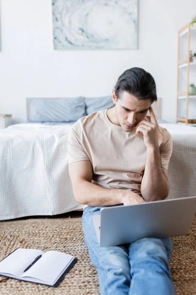 Pensive freelancer looking at laptop near notebook in bedroom — Stock Photo