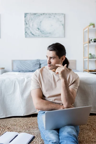 Pensive freelancer looking away near laptop and notebook in bedroom — Stock Photo