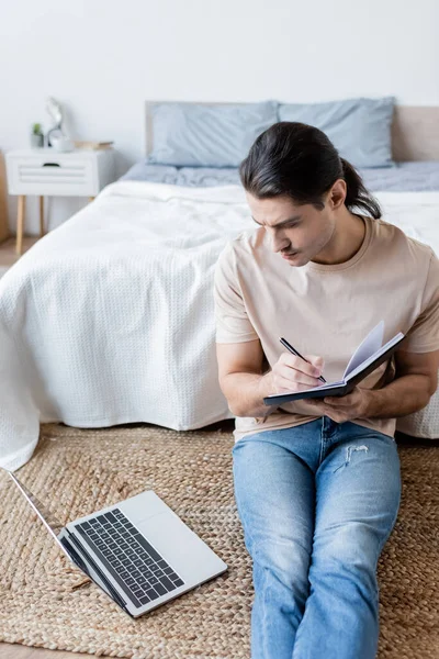Man with long hair writing in notebook and looking at laptop in bedroom — Stock Photo