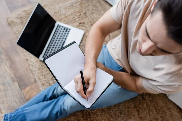 Top view of man writing in notebook near blurred laptop with blank screen — Stock Photo