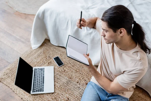 Mann mit langen Haaren hält Notizbuch und Stift in der Nähe von Laptop und Smartphone mit leerem Bildschirm im Schlafzimmer — Stockfoto