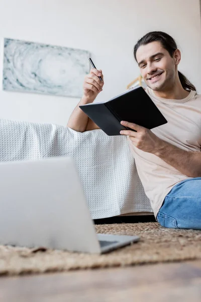 Hombre alegre con el pelo largo que sostiene el cuaderno y la pluma cerca del ordenador portátil borroso en el dormitorio - foto de stock