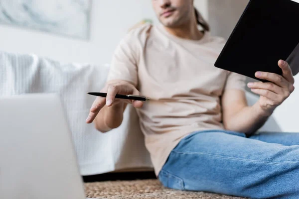 Ausgeschnittene Ansicht eines verschwommenen Mannes mit Notizbuch und Stift in der Nähe von Laptop im Schlafzimmer — Stockfoto