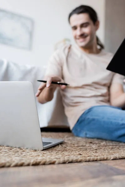 Homme flou tenant carnet et stylo près de l'ordinateur portable dans la chambre — Photo de stock