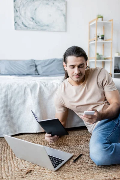 Man holding notebook and smartphone near laptop in bedroom — Stock Photo