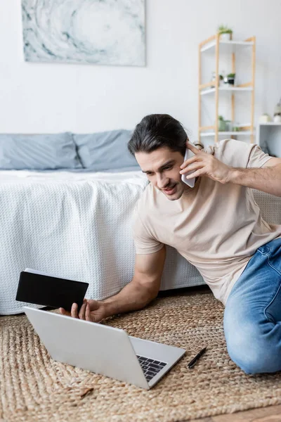 Мужчина держит ноутбук и разговаривает на смартфоне возле ноутбука в спальне — стоковое фото
