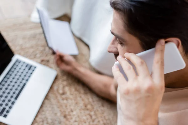 High angle view of man holding notebook and talking on smartphone near laptop — Stock Photo