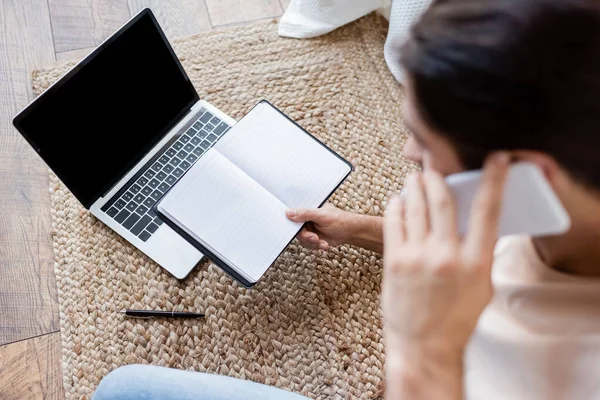 High angle view of blurred man holding notebook and talking on smartphone near laptop with blank screen — Stock Photo