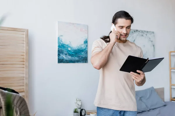 Hombre hablando por teléfono móvil y sosteniendo el cuaderno en casa - foto de stock