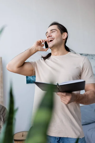 Uomo allegro parlando sul telefono cellulare e tenendo il taccuino a casa — Foto stock