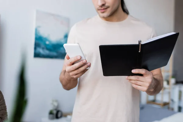 Cropped view of man holding mobile phone and notebook at home — Stock Photo