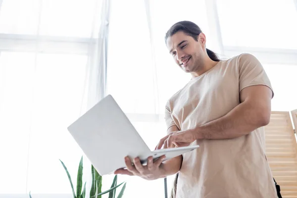 Baixo ângulo de visão do homem feliz com cabelos longos usando laptop — Fotografia de Stock