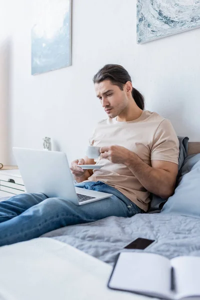 Man with long hair holding cup of coffee while using laptop in bedroom — Stock Photo