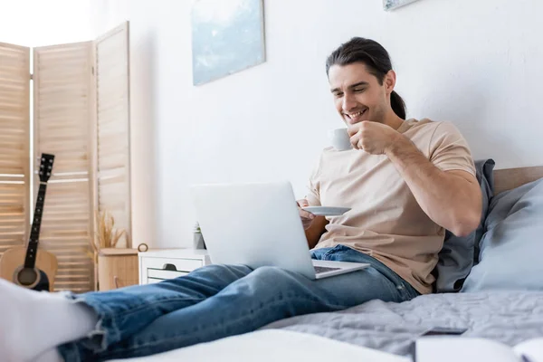 Cheerful man with long hair holding cup of coffee while using laptop in bedroom — Stock Photo