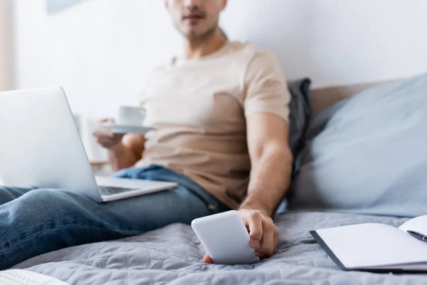 Cropped view of blurred man holding cup of coffee while reaching smartphone in bedroom — Stock Photo