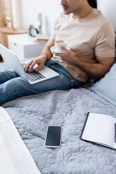Abgeschnittene Ansicht eines verschwommenen Mannes, der eine Tasse Kaffee hält, während er Laptop im Schlafzimmer benutzt — Stockfoto