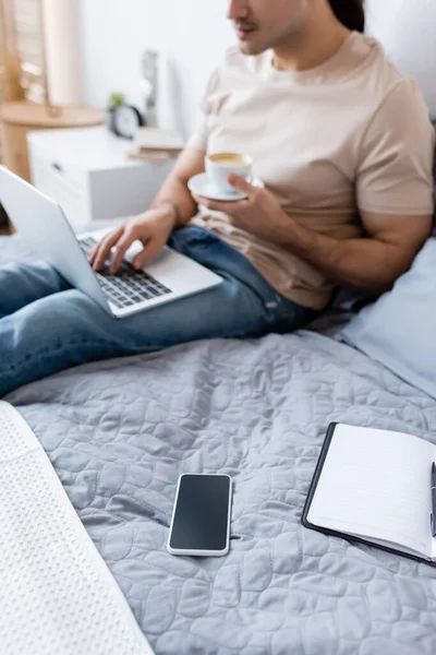 Vue recadrée de l'homme flou tenant tasse de café tout en utilisant un ordinateur portable dans la chambre — Photo de stock