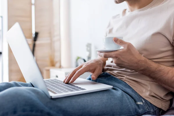 Cropped view of man holding cup of coffee while using laptop in bedroom — Stock Photo