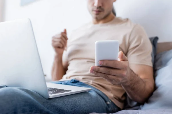 Cropped view of man holding smartphone near laptop in bedroom — Stock Photo
