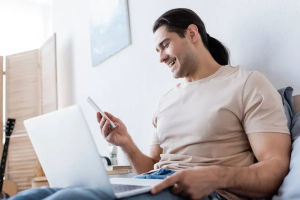 Fröhlicher Mann mit langen Haaren hält Smartphone neben Laptop im Schlafzimmer — Stockfoto