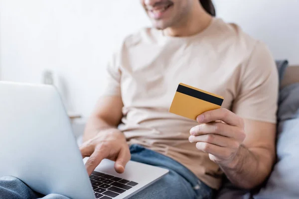 Vista ritagliata di felice uomo in possesso di carta di credito e utilizzando il computer portatile in camera da letto — Foto stock