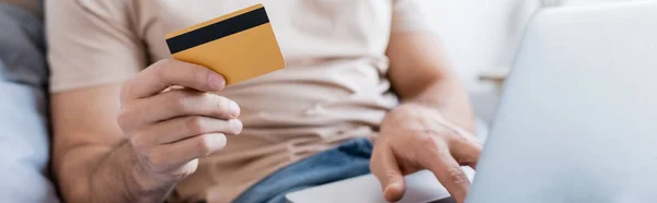 Cropped view of man holding credit card and using laptop in bedroom, banner — Stock Photo