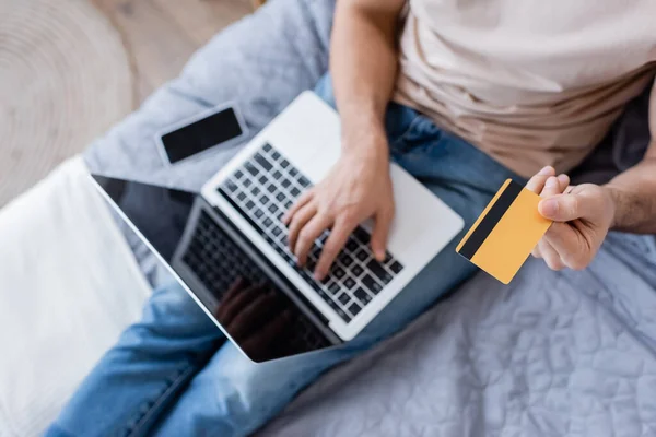 Vista superior del hombre que sostiene la tarjeta de crédito y el uso de un ordenador portátil cerca del teléfono inteligente en la cama - foto de stock