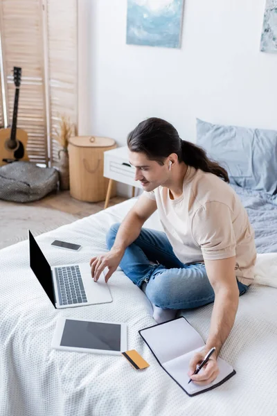 Smiling man with long hair in earphone writing in notebook and using laptop near gadgets on bed — Stock Photo