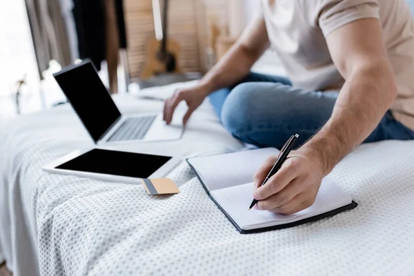 Vista cortada de homem escrevendo em notebook e usando laptop perto de cartão de crédito na cama — Fotografia de Stock