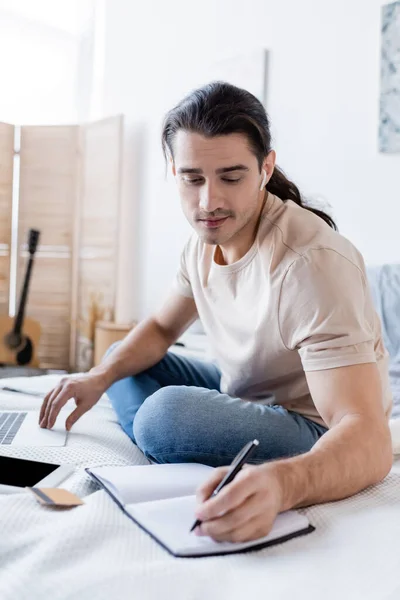 Uomo in auricolare scrivere in notebook vicino a carta di credito e gadget sul letto — Foto stock