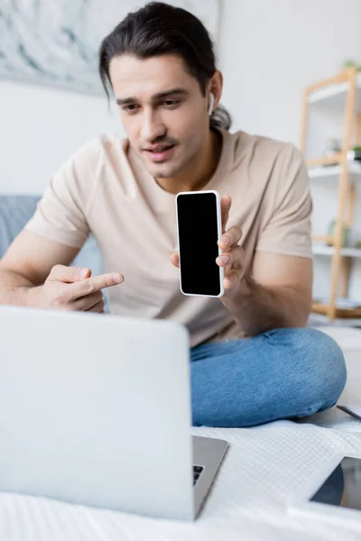 Man in earphone pointing at smartphone with blank screen during video call in bedroom — Stock Photo