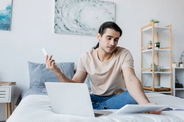 Homme dans l'écouteur tenant smartphone tout en regardant tablette numérique près d'un ordinateur portable sur le lit — Photo de stock