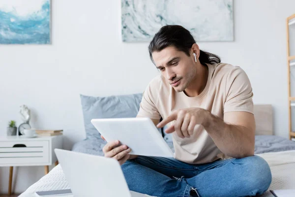 Homem em fone de ouvido apontando para tablet digital perto de laptop e smartphone na cama — Fotografia de Stock