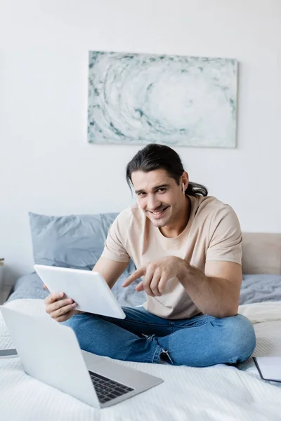 Joyful man in earphone pointing at digital tablet near laptop and smartphone on bed — Stock Photo