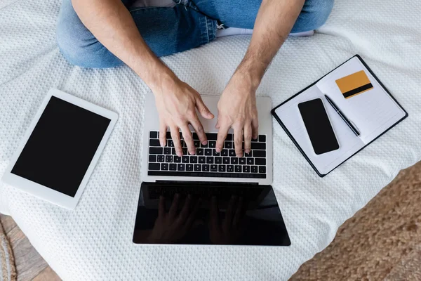 Vista cortada do homem usando laptop com tela em branco perto de gadgets e cartão de crédito na cama — Fotografia de Stock