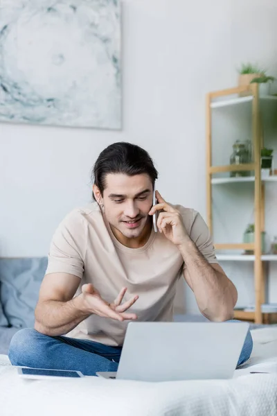 Homme souriant parlant sur un téléphone mobile tout en gesticulant près d'un ordinateur portable dans la chambre — Photo de stock