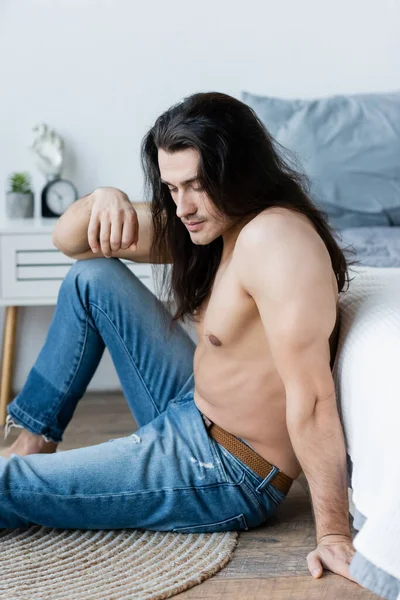 Hombre sin camisa pensativo con el pelo largo sentado cerca de la cama - foto de stock